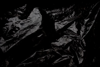 Crumpled and glossy black material with a texture resembling plastic or fabric. The surface displays various folds and creases, creating an abstract pattern.