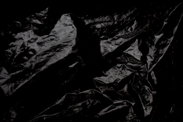 Crumpled and glossy black material with a texture resembling plastic or fabric. The surface displays various folds and creases, creating an abstract pattern.