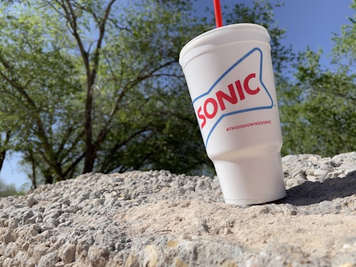 A large white cup with the Sonic logo is placed on a textured stone surface. In the background, there are green trees against a clear blue sky.