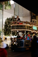 A festive tram decorated with holiday lights and ornaments is moving along a street. A person dressed as Santa Claus is standing on the top deck, waving. The tram is adorned with garlands and bows, contributing to the cheerful holiday atmosphere. Trees with string lights are visible in the background, suggesting a shopping area with glass storefronts.
