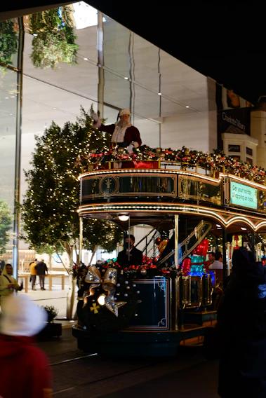 A festive tram decorated with holiday lights and ornaments is moving along a street. A person dressed as Santa Claus is standing on the top deck, waving. The tram is adorned with garlands and bows, contributing to the cheerful holiday atmosphere. Trees with string lights are visible in the background, suggesting a shopping area with glass storefronts.