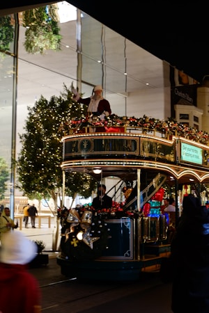 A festive tram decorated with holiday lights and ornaments is moving along a street. A person dressed as Santa Claus is standing on the top deck, waving. The tram is adorned with garlands and bows, contributing to the cheerful holiday atmosphere. Trees with string lights are visible in the background, suggesting a shopping area with glass storefronts.