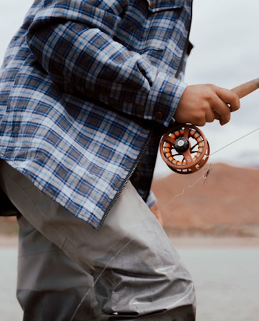 A person wearing a plaid shirt and waterproof waders is holding a fishing rod with a fly reel. The focus is on the hand gripping the rod and the attire. The background is slightly blurred, suggesting an outdoor setting near water.
