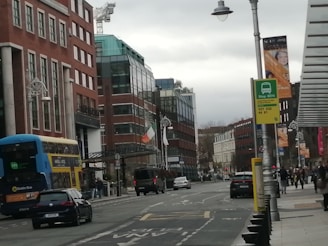 A city street with modern buildings on either side, featuring a mix of glass and brick. A double-decker bus is visible on the left, along with several cars driving along the road. A prominent green and orange flag is flying on the right. There are street lamps and signs for a bus stop displaying route numbers. Sparse foot traffic is seen with a few pedestrians on the sidewalk.