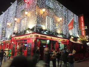 A bustling evening scene captures a lively bar adorned with bright red facades and illuminated with festive string lights. People gather outside, some in motion, contributing to a dynamic and vibrant atmosphere. The bar is decorated with traditional signage and greenery.