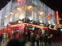 A bustling evening scene captures a lively bar adorned with bright red facades and illuminated with festive string lights. People gather outside, some in motion, contributing to a dynamic and vibrant atmosphere. The bar is decorated with traditional signage and greenery.
