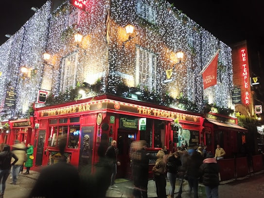 A bustling evening scene captures a lively bar adorned with bright red facades and illuminated with festive string lights. People gather outside, some in motion, contributing to a dynamic and vibrant atmosphere. The bar is decorated with traditional signage and greenery.