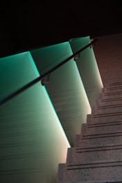 A modern staircase with a sleek glass railing illuminated by soft teal lighting creates a visually striking effect. The stepped shadows and speckled surfaces add texture and depth, contributing to a minimalist design aesthetic.