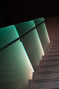 A modern staircase with a sleek glass railing illuminated by soft teal lighting creates a visually striking effect. The stepped shadows and speckled surfaces add texture and depth, contributing to a minimalist design aesthetic.