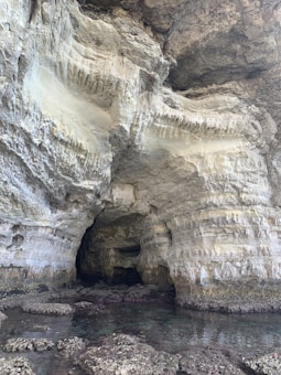 A natural cave with rugged, textured rock formations featuring various shades of beige and gray. The entrance of the cave leads into a darker interior, while shallow water with visible rocks appears in the foreground.