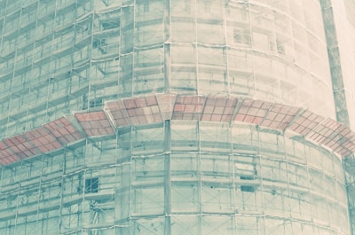 A large building covered in scaffolding and protective netting. The structure is partially visible through the translucent material, with metal beams and construction platforms encircling the exterior.