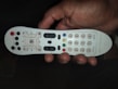 A person's hand is holding a white remote control that has various buttons in black and colored in red, blue, green, and yellow. There are numeric keys on the right side while the left side contains function buttons such as power, menu, and volume controls.