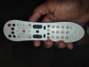 A person's hand is holding a white remote control that has various buttons in black and colored in red, blue, green, and yellow. There are numeric keys on the right side while the left side contains function buttons such as power, menu, and volume controls.
