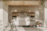 A well-organized and aesthetically designed bar area features a neatly arranged collection of liquor bottles and bar equipment. The countertop is clean, with several metal shakers, glassware, and a few plates with food items like bread or pastries. Potted plants hang elegantly from the ceiling, casting shadows on the light-tiled surfaces and walls, giving the space a natural and inviting ambience.