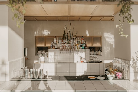 A well-organized and aesthetically designed bar area features a neatly arranged collection of liquor bottles and bar equipment. The countertop is clean, with several metal shakers, glassware, and a few plates with food items like bread or pastries. Potted plants hang elegantly from the ceiling, casting shadows on the light-tiled surfaces and walls, giving the space a natural and inviting ambience.