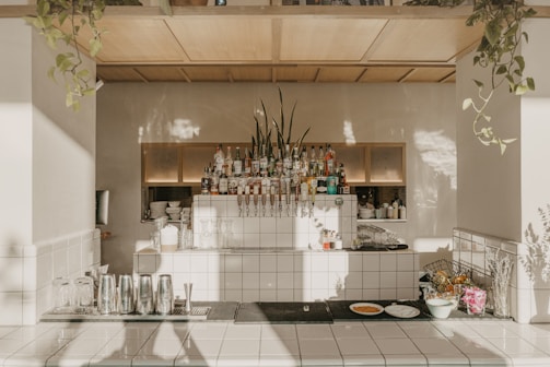 A well-organized and aesthetically designed bar area features a neatly arranged collection of liquor bottles and bar equipment. The countertop is clean, with several metal shakers, glassware, and a few plates with food items like bread or pastries. Potted plants hang elegantly from the ceiling, casting shadows on the light-tiled surfaces and walls, giving the space a natural and inviting ambience.