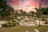 A beautifully landscaped garden featuring neatly trimmed grass, palm trees, and curving stone pathways. A vibrant sunset with pink, orange, and purple hues fills the sky, providing a colorful backdrop. The garden's layout includes various circular and spiral patterns, bordered by low hedges and decorative plants.