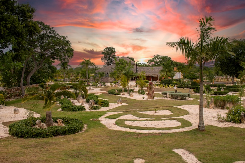 A beautifully landscaped garden featuring neatly trimmed grass, palm trees, and curving stone pathways. A vibrant sunset with pink, orange, and purple hues fills the sky, providing a colorful backdrop. The garden's layout includes various circular and spiral patterns, bordered by low hedges and decorative plants.