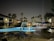 A nighttime scene of a serene outdoor swimming pool surrounded by lounge chairs with blue fabric and large umbrellas. Palm trees are prominently visible around the area, set against a backdrop of residential buildings. The pool is illuminated, reflecting a bluish glow on the wet poolside area.