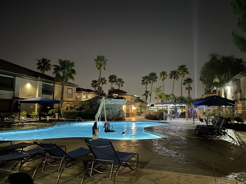 A nighttime scene of a serene outdoor swimming pool surrounded by lounge chairs with blue fabric and large umbrellas. Palm trees are prominently visible around the area, set against a backdrop of residential buildings. The pool is illuminated, reflecting a bluish glow on the wet poolside area.