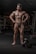 A muscular man is posing in a gym, standing confidently with hands on his hips. The gym equipment is visible in the background, and a dumbbell is lying on the floor near his feet. The lighting highlights the man's muscles, giving emphasis to his physique.