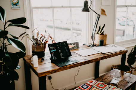A cozy home office desk with a laptop, ledger books, and a cup of tea by a window overlooking the Cotswold countryside.