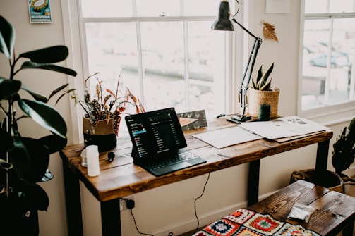 A cozy home office or study space featuring a wooden desk with a laptop, papers, and a desk lamp. Several potted plants add greenery to the room. A large window lets in natural light, and a bright crocheted cushion is on the bench next to the desk.