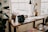 A cozy home office or study space featuring a wooden desk with a laptop, papers, and a desk lamp. Several potted plants add greenery to the room. A large window lets in natural light, and a bright crocheted cushion is on the bench next to the desk.