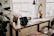 A cozy home office or study space featuring a wooden desk with a laptop, papers, and a desk lamp. Several potted plants add greenery to the room. A large window lets in natural light, and a bright crocheted cushion is on the bench next to the desk.