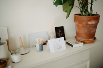 A cozy interior space features a tabletop arrangement with various items. There is a tall terracotta pot containing a green plant, a candle placed on a ceramic holder, a small jar filled with matches, and a selection of cards or photographs standing upright. The overall atmosphere is artistic and serene.