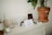 A cozy interior space features a tabletop arrangement with various items. There is a tall terracotta pot containing a green plant, a candle placed on a ceramic holder, a small jar filled with matches, and a selection of cards or photographs standing upright. The overall atmosphere is artistic and serene.