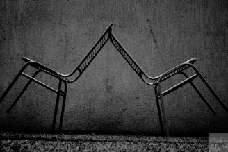Two metal chairs are positioned in a balancing act with their backrests touching, creating an angular, symmetrical form against a textured concrete wall. The scene is captured in black and white, enhancing the contrast between the dark chairs and the lighter background.