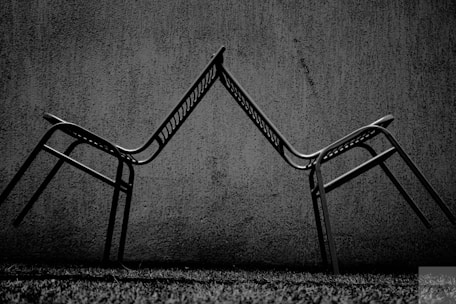 Two metal chairs are positioned in a balancing act with their backrests touching, creating an angular, symmetrical form against a textured concrete wall. The scene is captured in black and white, enhancing the contrast between the dark chairs and the lighter background.