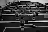 A black and white image of a foosball table with multiple rows of small, plastic soccer player figures mounted on rods. The figures are wearing sports uniforms and are positioned over the table's pitch. The surface of the table displays circular patterns resembling a soccer field.