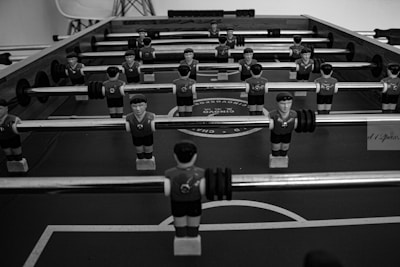 A black and white image of a foosball table with multiple rows of small, plastic soccer player figures mounted on rods. The figures are wearing sports uniforms and are positioned over the table's pitch. The surface of the table displays circular patterns resembling a soccer field.