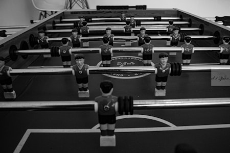 A black and white image of a foosball table with multiple rows of small, plastic soccer player figures mounted on rods. The figures are wearing sports uniforms and are positioned over the table's pitch. The surface of the table displays circular patterns resembling a soccer field.