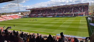 A large football stadium filled with spectators, with a well-maintained green field and a match in progress. The stands are partially covered and packed with fans. Clear skies and a few clouds can be seen above the large, modern stadium structure.
