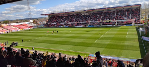 A large football stadium filled with spectators, with a well-maintained green field and a match in progress. The stands are partially covered and packed with fans. Clear skies and a few clouds can be seen above the large, modern stadium structure.