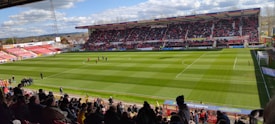 A large football stadium filled with spectators, with a well-maintained green field and a match in progress. The stands are partially covered and packed with fans. Clear skies and a few clouds can be seen above the large, modern stadium structure.