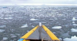 A kayak is navigating through a vast expanse of icy water with small ice floes scattered across the surface. The sky is overcast, casting a gray tone over the scene. The yellow front of the kayak adds a pop of color against the cold, blue-gray palette.