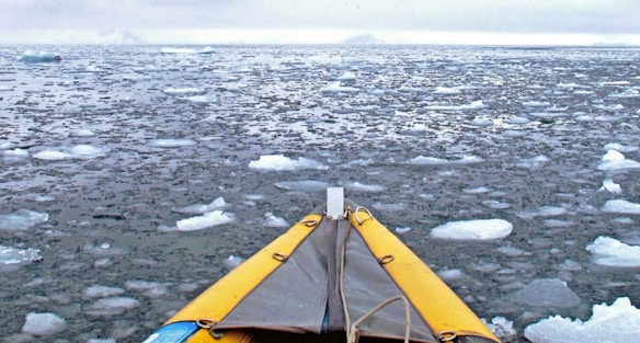 A kayak is navigating through a vast expanse of icy water with small ice floes scattered across the surface. The sky is overcast, casting a gray tone over the scene. The yellow front of the kayak adds a pop of color against the cold, blue-gray palette.