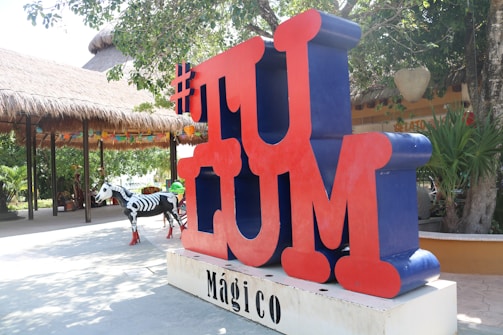 Large red and blue 3D letters spell out 'TULUM' with a hashtag symbol at the top. The background shows a thatched-roof structure and lush greenery, while a decorative zebra painted with skeletal patterns stands nearby.