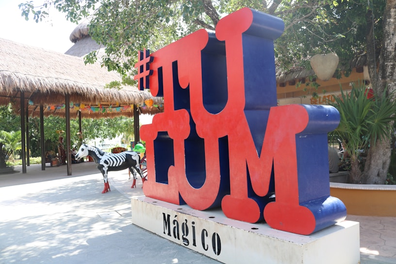 Large red and blue 3D letters spell out 'TULUM' with a hashtag symbol at the top. The background shows a thatched-roof structure and lush greenery, while a decorative zebra painted with skeletal patterns stands nearby.