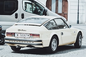 A vintage Alfa Romeo car is parked on a cobblestone street, showcasing its retro design and cream color. Nearby, a modern van and another vehicle are partially visible. The setting appears to be urban with old-style building facades and arched wooden doors in the background.