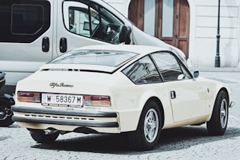 A vintage Alfa Romeo car is parked on a cobblestone street, showcasing its retro design and cream color. Nearby, a modern van and another vehicle are partially visible. The setting appears to be urban with old-style building facades and arched wooden doors in the background.