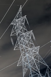 An inverted image of a large electricity transmission tower made of metal. The intricate lattice structure stands out sharply against a dark, cloudy background.