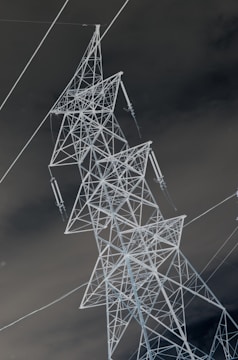 An inverted image of a large electricity transmission tower made of metal. The intricate lattice structure stands out sharply against a dark, cloudy background.