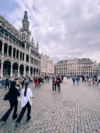 A historic European square with ornate buildings featuring intricate architectural details and spires. The wide cobblestone plaza is bustling with people walking and gathering, under a cloudy sky. The atmosphere is lively with a mix of tourists and locals.