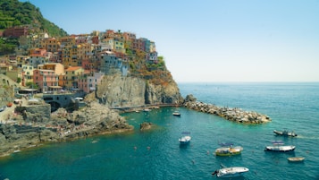 A coastal village with colorful buildings perched on a rocky cliff overlooking the sea. The water is a vibrant blue-green, and several small boats float in the harbor. A stone walkway and pier extend into the water, with people walking and sitting nearby. The landscape is lush with greenery and the sky is clear and bright.