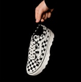 A hand holding a black and white checkerboard patterned sneaker against a solid black background. The sneaker features a thick white sole and white laces, with a classic lace-up design. The minimalist setting emphasizes the shoe's bold pattern.